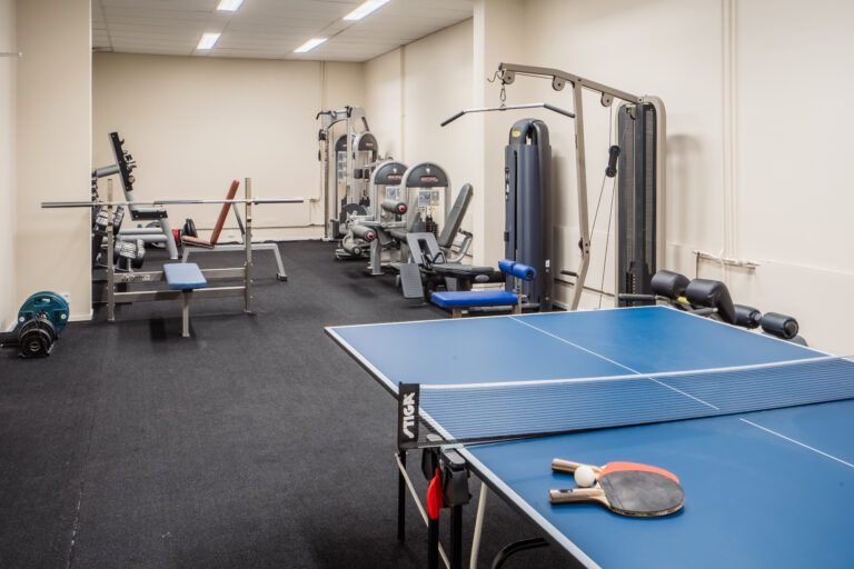 The machine area of a student housing gym with multiple exercise machines and a nearby table tennis table for residents.