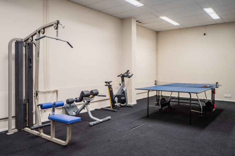 A view of the student housing gym with various workout machines and a ping pong table in a spacious room.